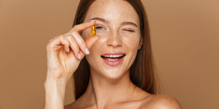 Beauty portrait of a beautiful healthy young topless woman with long red hair showing vitamin capsule isolated over beige background